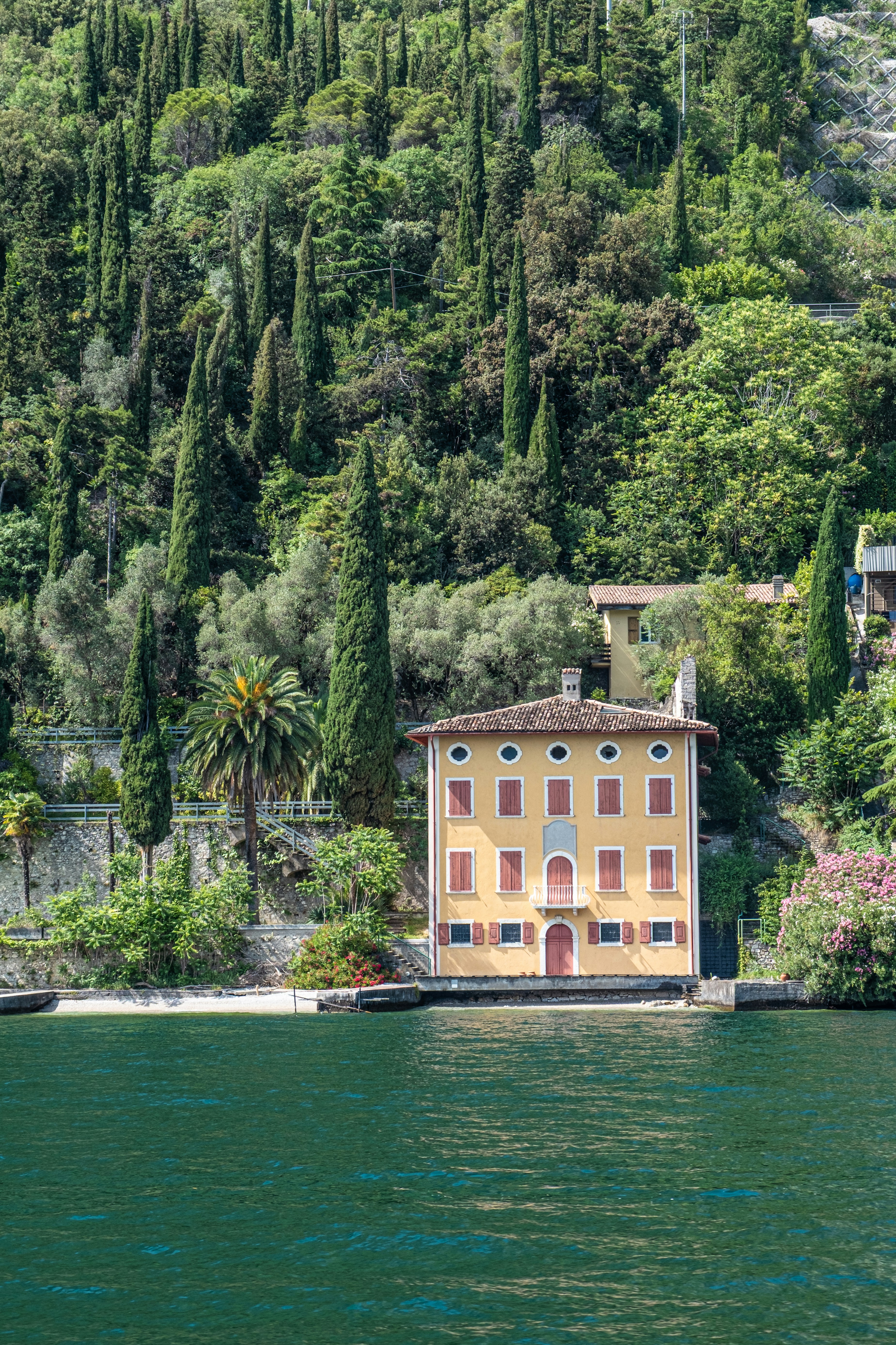 A warm ochre lakeside villa surrounded by cypress and olive trees, with a palm and green water